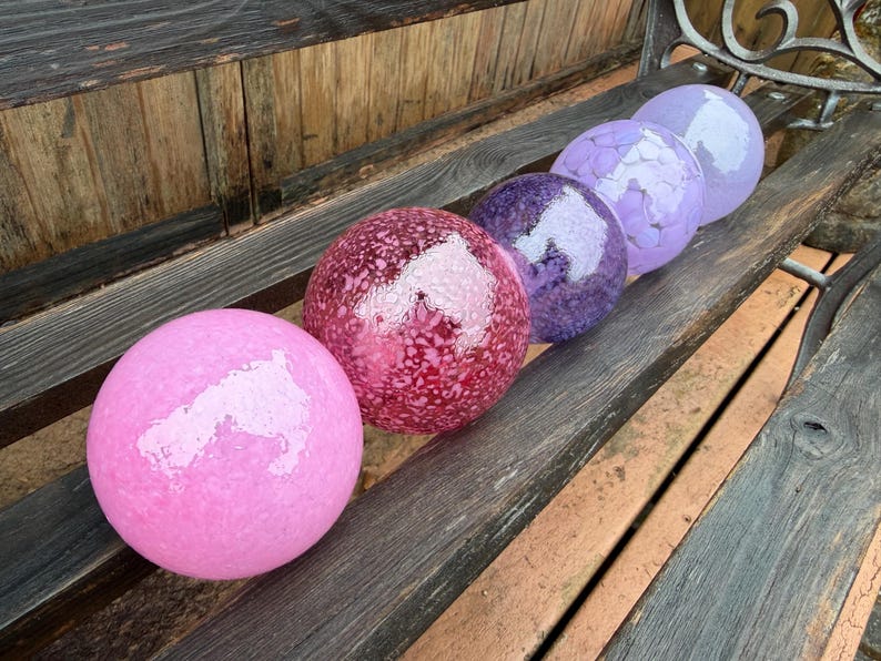 May include: Six decorative glass spheres in shades of pink, red, purple, and lavender are arranged on a wooden bench. The spheres have textured surfaces and reflect light, creating a visually appealing display. The spheres vary in size and color.