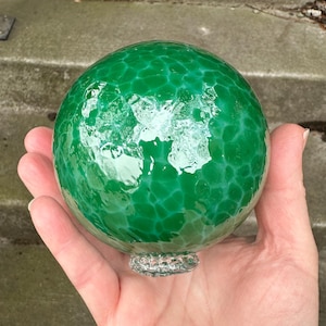 May include: A green glass ball with a marbled pattern. The ball is smooth and shiny.