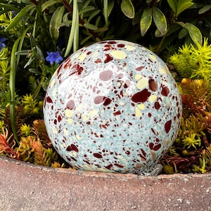May include: A large, round glass orb with a mottled blue, white, and brown pattern. The orb is sitting on a bed of green and brown foliage.