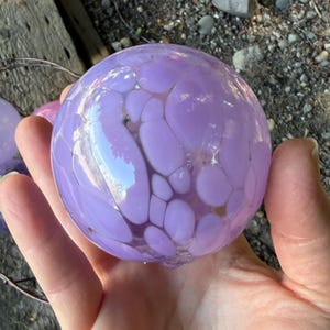 May include: A hand holds a large, round, lavender-colored glass orb with a speckled pattern. The orb is reflective, with lighter purple spots. Other smaller orbs are visible in the background, along with a metal wire basket.