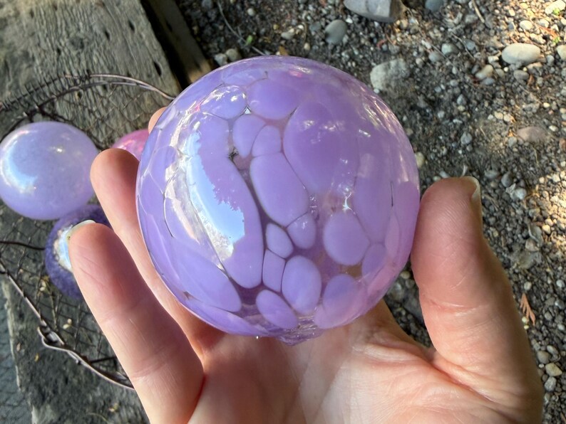 May include: A hand holds a large, round, lavender-colored glass orb with a speckled pattern. The orb is reflective, with lighter purple spots. Other smaller orbs are visible in the background, along with a metal wire basket.