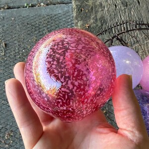 May include: A collection of decorative glass spheres in various shades of pink, purple, and blue. The spheres have a speckled pattern and are held in a hand. The spheres are in a wire basket.