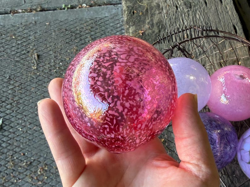 May include: A collection of decorative glass spheres in various shades of pink, purple, and blue. The spheres have a speckled pattern and are held in a hand. The spheres are in a wire basket.