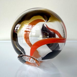 May include: A spherical glass vase featuring clear glass with swirling ribbons of deep brown, black, and vibrant orange. The piece has a small, circular clear glass base that supports the rounded body, creating a smooth, reflective surface.