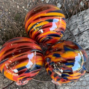 May include: Three spherical glass ornaments featuring a swirling pattern of vibrant orange, deep red, and dark blue colors. Each smooth, polished orb has a glossy finish that reflects light, showcasing a marbled effect throughout the glass surface.