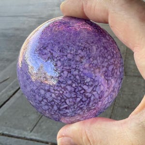 May include: A round, purple glass orb with a textured surface. The orb is held in a hand and reflects the surrounding environment. The color is a mix of light and dark purples.