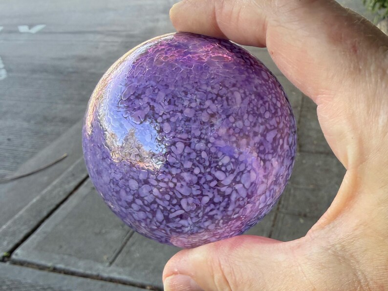 May include: A round, purple glass orb with a textured surface. The orb is held in a hand and reflects the surrounding environment. The color is a mix of light and dark purples.