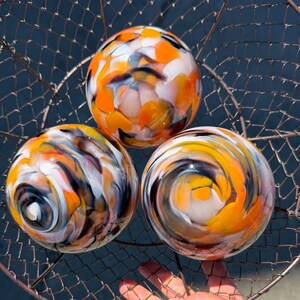 May include: Three spherical glass ornaments featuring a swirling pattern of orange, yellow, white, and dark gray colors. The smooth, polished glass spheres are arranged together inside a wire mesh basket with a metallic finish.