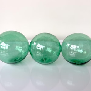 May include: Three spherical glass fishing floats in a translucent light green color. Each orb features a smooth, glossy surface with subtle internal air bubbles and a small, circular indentation at the base, reflecting light across their rounded forms.