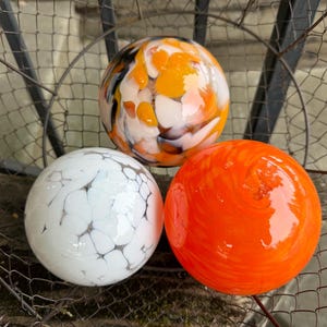 May include: Three decorative glass spheres in a metal basket. One sphere is white with black veining, another is bright orange, and the third is white with orange and black swirls. The spheres are round and have a glossy finish.