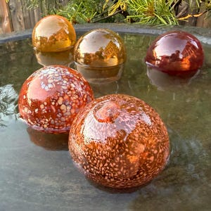 May include: Five glass spheres floating in water. Three spheres are translucent in shades of amber and deep red. Two spheres in the foreground feature a speckled, multi-colored pattern with white, orange, and brown flecks on a reddish-brown base.