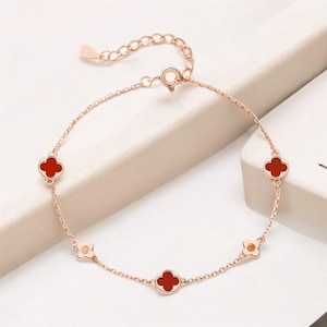 May include: Rose gold bracelet featuring four-leaf clover charms with red centers. The delicate chain is adjustable and has a lobster clasp closure. The bracelet is a stylish accessory.