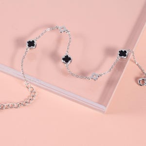 May include: A silver bracelet with a delicate chain design. The bracelet features four-leaf clover charms with black centers and a small, adjustable chain. The bracelet is displayed on a pink surface.