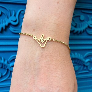 May include: Gold-colored bracelet featuring a 'shaka' hand gesture charm. The charm is a cutout design, and the bracelet has a delicate chain. The bracelet is worn on a wrist, against a blue background.