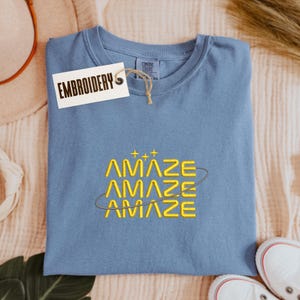 Może przedstawiać: Niebieska koszulka Comfort Colors z wyhaftowanym słowem "AMAZE" żółtą nicią. Słowo jest powtórzone trzy razy ze stylizowanym wzorem orbity. Do koszulki dołączona jest metka z napisem "EMBROIDERY".