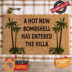 May include: A rectangular tan coir doormat featuring black text that reads A HOT NEW BOMBSHELL HAS ENTERED THE VILLA. The design includes two green palm tree illustrations, one on the left side and one on the right side of the central text.