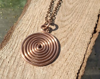 May include: A copper-colored metal pendant shaped as a flat, circular spiral. The pendant is attached to a matching copper-colored metal chain link necklace. The design features a single continuous wire coiled tightly into a round, flat shape.