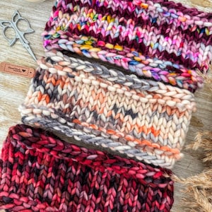 May include: Three chunky knit headbands in various colours. The top headband is a mix of pinks, purples, and yellows. The middle one is cream, grey, and orange. The bottom one is a deep red. A lit candle and scissors are in the background.