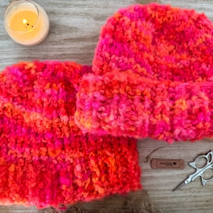 May include: A vibrant, hand-knitted hat and scarf set in shades of pink and orange. The textured knitwear is displayed on a wooden surface, alongside a lit candle, a small tag, and decorative scissors. The set is a cozy accessory.