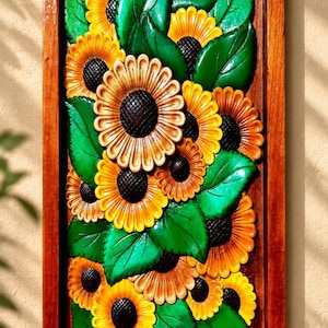Puede incluir: Una pieza de arte mural rectangular de madera que presenta una talla en alto relieve de varios girasoles con pétalos amarillos y naranjas, centros de color marrón oscuro y hojas de color verde vibrante, enmarcada en madera marrón pulida con el texto Ibu Yuma DESIGN.