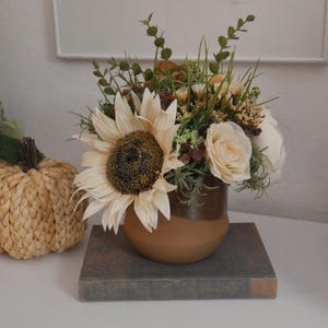 Puede incluir: Arreglo floral decorativo con un gran girasol color crema y otras flores en un jarrón de cerámica marrón. También se ven una calabaza tejida y un libro. Para la decoración del hogar.