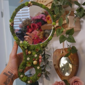May include: A round hand mirror with a green moss-covered frame and handle, adorned with small, decorative elements. The mirror reflects a colorful floral arrangement. The handle has a unique, organic shape, adding to its whimsical appeal.