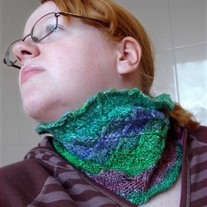 Puede incluir: Un cuello tejido a mano en tonos verdes, azules y morados. El cuello tiene un borde con volantes y se lleva sobre una camisa a rayas marrones y moradas. La textura tejida es visible.