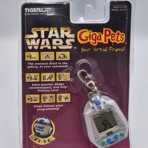 Può includere: Un giocattolo elettronico Star Wars Giga Pets. La confezione presenta il logo di Star Wars e il testo "Giga Pets, Your Virtual Friend!". Il giocattolo ha la forma di R2-D2 e ha un piccolo schermo e pulsanti.