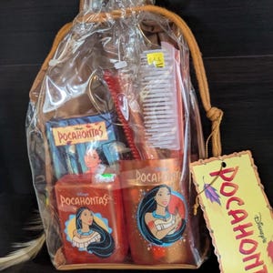 Può includere: Un sacchetto regalo trasparente contenente oggetti a tema Pocahontas. La borsa include un pettine, una spazzola e due contenitori con l'immagine di Pocahontas. Un'etichetta gialla con il nome "Pocahontas" e il logo Disney è attaccata.