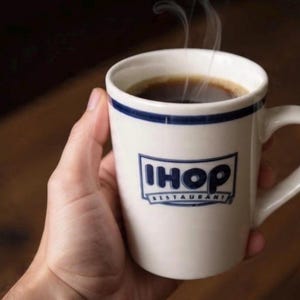 May include: A white IHOP restaurant coffee mug with a blue rim and logo, filled with hot coffee. Steam rises from the dark liquid. The mug is held in a hand.