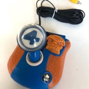 May include: A vintage Fantastic Four video game controller. The controller is orange and blue, featuring a clear button with the number 4 and a character likeness. It has a black cable with a yellow connector.