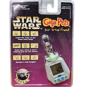 Può includere: Un giocattolo elettronico per animali virtuali Star Wars Giga Pets, confezionato in un blister di plastica trasparente. Il giocattolo presenta un piccolo schermo e pulsanti, con un personaggio di Yoda attaccato. La confezione include il testo "Impara le vie della Forza da Yoda!"