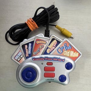 Puede incluir: Una consola de juegos Family Fun 10 en 1 de color plateado con un cable negro y un conector RCA amarillo. La consola tiene un joystick y botones azules, con títulos de juegos como "Hang Man" y "Go Fish".