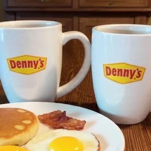 May include: Two white mugs with the Denny's logo, a plate with a pancake, bacon, and two fried eggs. The mugs are on a wooden table. One hand is holding one of the mugs.