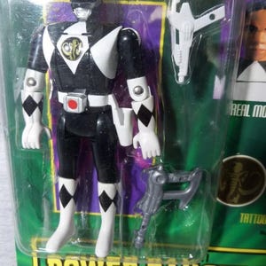 May include: A Power Rangers action figure in a sealed package. The figure is black and white with a helmet, gloves, and boots. It has a gold emblem on the chest and comes with two accessories. The packaging has the words "Power Rangers" in a green and yellow font.
