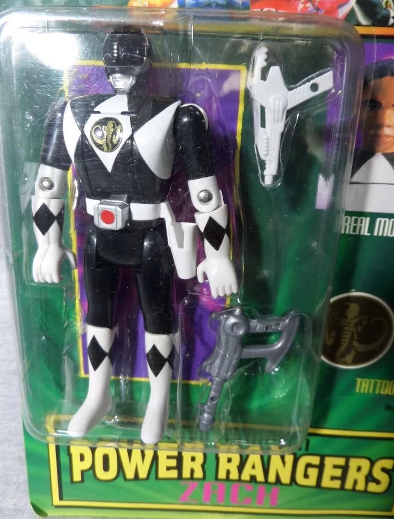 May include: A Power Rangers action figure in a sealed package. The figure is black and white with a helmet, gloves, and boots. It has a gold emblem on the chest and comes with two accessories. The packaging has the words "Power Rangers" in a green and yellow font.
