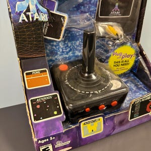 以下が含まれることがあります： 黒いジョイスティックと赤いボタンが付いたAtariビデオゲームシステム。パッケージには、Centipede、Asteroids、Pong、Adventureのゲームスクリーンショットが含まれています。箱には「Plug and play! This is all you need!」と「Ages 5+」と書かれています。