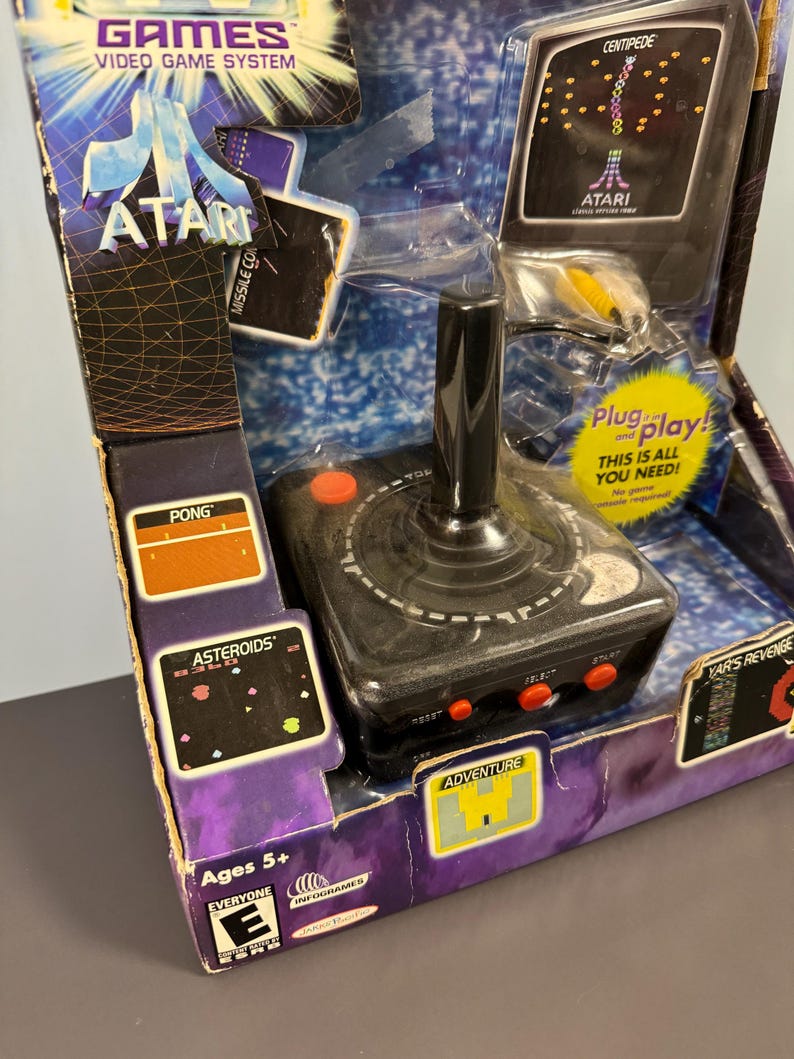以下が含まれることがあります： 黒いジョイスティックと赤いボタンが付いたAtariビデオゲームシステム。パッケージには、Centipede、Asteroids、Pong、Adventureのゲームスクリーンショットが含まれています。箱には「Plug and play! This is all you need!」と「Ages 5+」と書かれています。
