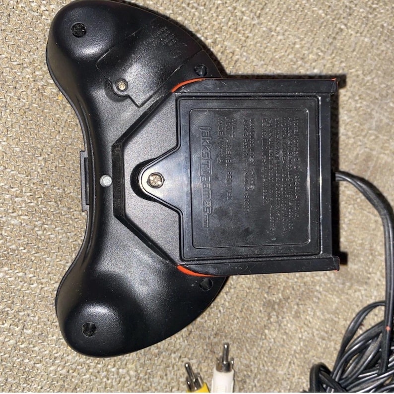 May include: A black plastic video game controller with a rectangular battery compartment on the back. The device features the Jakks Pacific logo and text molded into the casing, along with a black wired cable ending in yellow and white RCA plugs.
