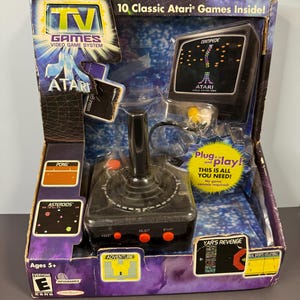 以下が含まれることがあります： 箱入りのAtari TV Gamesビデオゲームシステム。パッケージは青と紫で、Atariのロゴと「Centipede」や「Yar's Revenge」などのゲームタイトルが見えます。赤いボタンと小さな画面が付いた黒いジョイスティックが含まれています。