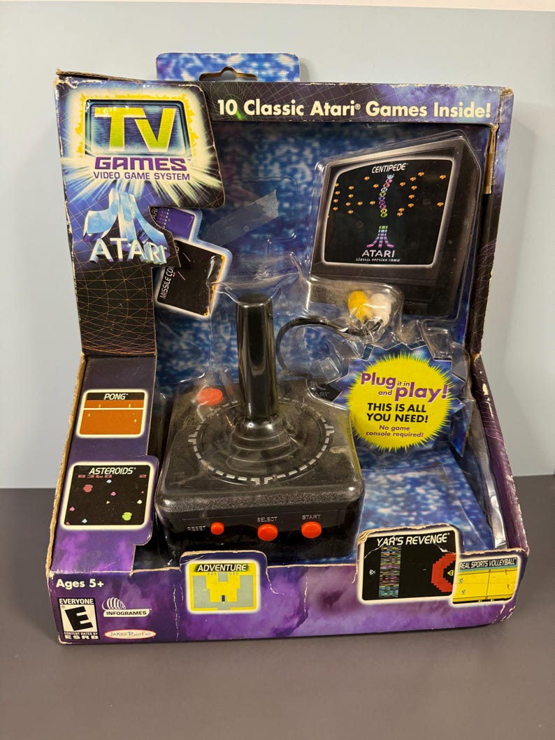 以下が含まれることがあります： 箱入りのAtari TV Gamesビデオゲームシステム。パッケージは青と紫で、Atariのロゴと「Centipede」や「Yar's Revenge」などのゲームタイトルが見えます。赤いボタンと小さな画面が付いた黒いジョイスティックが含まれています。