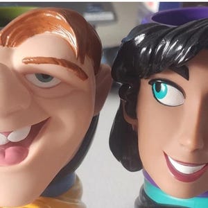 May include: Two ceramic mugs featuring cartoon character faces. One mug depicts a man with orange hair and a large nose, the other depicts a man with black hair and teal eyes. The mugs have a purple and green interior.