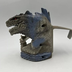May include: A Godzilla-themed toy in shades of gray and blue. The toy features a detailed dinosaur head with red eyes and an open mouth, along with a handle on the side. The toy is designed to resemble the iconic movie monster.