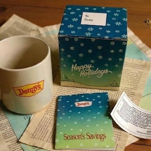 May include: A beige Denny's mug, a gift box, and a small booklet are displayed on a newspaper background. The mug has the Denny's logo. The gift box is blue with white snowflake designs and the words "Happy Holidays". The booklet says "Season's Savings".
