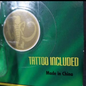 May include: A gold-colored circular emblem with an elephant design, set against a green background. The text "TATTOO INCLUDED" is printed in yellow. The words "Made in China" are also visible.