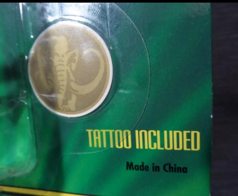 May include: A gold-colored circular emblem with an elephant design, set against a green background. The text "TATTOO INCLUDED" is printed in yellow. The words "Made in China" are also visible.