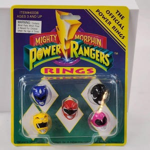 Può includere: Un pacchetto di anelli Mighty Morphin Power Rangers. La confezione è gialla e verde e presenta il logo Power Rangers. Gli anelli sono in un vassoio di plastica trasparente e sono rossi, blu, gialli, rosa e neri.