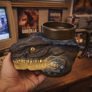 Può includere: Tazza a tema Godzilla, con una testa scolpita dettagliata, occhi gialli e un manico marrone. La tazza è blu scuro e marrone, con una parte superiore rotonda per contenere liquidi. Il design è ispirato all'iconico mostro cinematografico.