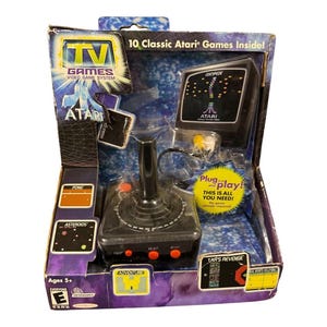 以下が含まれることがあります： 箱入りのAtari TV Gamesビデオゲームシステム。パッケージには、Atariのロゴ、ゲームタイトル、ジョイスティックコントローラーが描かれています。箱には「10 Classic Atari Games Inside!」と「Plug and play!」の文字が含まれています。
