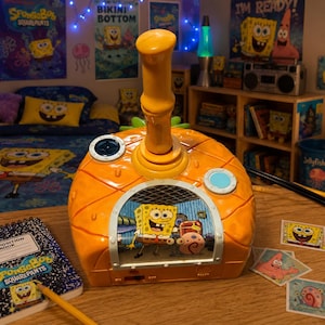 Puede incluir: Una casa de juguete de plástico naranja brillante con forma de piña que presenta una ventana central con Bob Esponja y Gary el caracol. La estructura incluye una chimenea alta, un ojo de buey azul circular y un botón circular oscuro arriba.